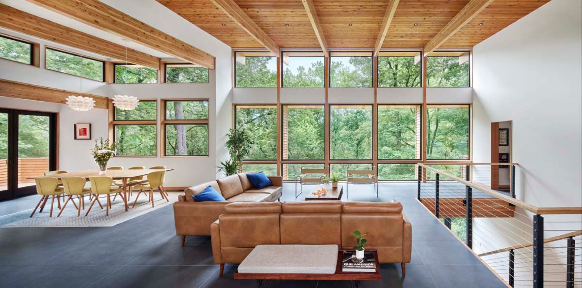 Open concept MCM living/dining room with wood beams & forest views through floor to ceiling black metal frame windows.